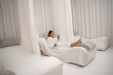 Beyaz bornozlu mutlu kadın spa salonunda bir fincan kahveyle eğleniyor.