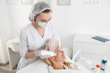 Kadın kozmetik uzmanı, modern klinikte ultrasonik yüz temizleme yöntemi kullanarak kadın hasta ile çalışıyor.