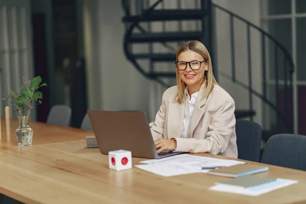 現代のオフィスに座っている間 ラップトップで働く大人のビジネス女性を笑顔 ぼやけた背景 — ストック写真