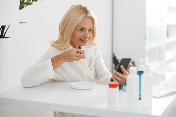 白人熟女はカップを保持し 現代のスパセンターでスマートフォンを見て 美容クリニック — ストック写真