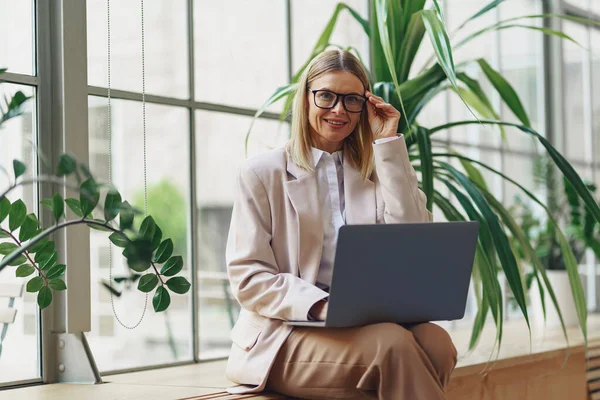 坐在现代办公室的同时 微笑着工作在笔记本电脑上的成年女商人 模糊的背景 — 图库照片