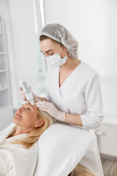 プロの若い女性美容師作る超音波顔剥離のために幸せな白人成熟したかなり女性患者でキャビネット — ストック写真