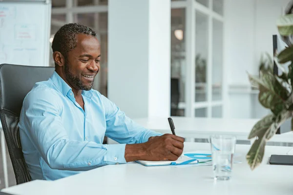 現代のオフィスに座っている間にメモを取るアフリカのビジネスマンの笑顔 高品質の写真 — ストック写真