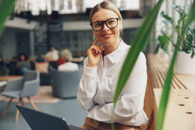 Pencerenin yanındaki ofiste dizüstü bilgisayarla çalışan şık bir kadın kameraya bakıyor. Yüksek kalite fotoğraf