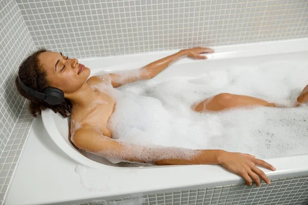 stock image Beautiful African woman in headphone takes bubble bath in bathtub and relaxing with closed eyes