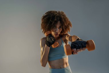 Kaslı bir kadın stüdyo arka planında ağırlık kaldırarak egzersiz yapıyor ve kameraya bakıyor.