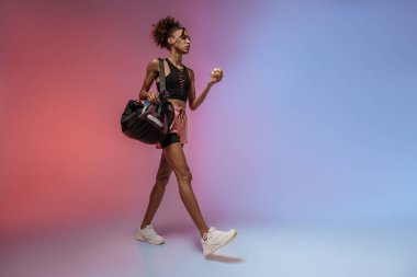 Stüdyo arka planında su şişesi, spor çantası ve elma taşıyan bir kadın antrenmana gidiyor.