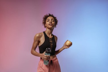 Stüdyo arka planında çalıştıktan sonra elinde bir şişe su ve elma tutan yorgun bir kadın.