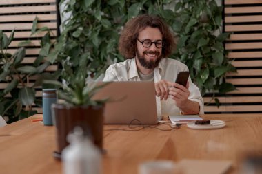 Dizüstü bilgisayarda çalışan serbest çalışan bir erkek ve modern iş yerinde otururken telefon kullanıyor.