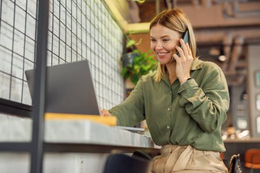 Gülümseyen iş kadını iş yerinde çalışırken telefonla konuşuyor ve bilgisayarda çalışıyor.