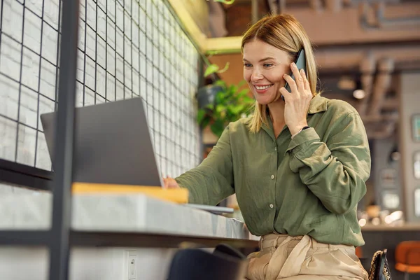 stock image Smiling businesswoman is talking phone while sitting in coworking and work on laptop