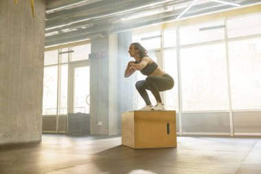 Spor salonundaki tahta kutunun üzerinde duran atletik kadın çömeliyor. Yüksek kalite fotoğraf