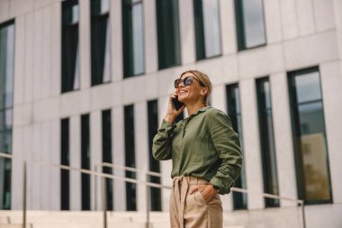 Smiling woman office worker is talking by phone while standing on modern building background 