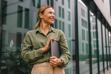 Gülümseyen kadın ofis çalışanı modern bina arka planında duruyor ve yan tarafa bakıyor.