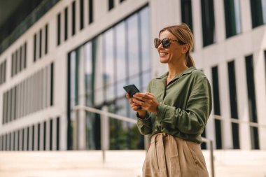 Smiling woman office worker in sunglasses is standing on modern building background with phone