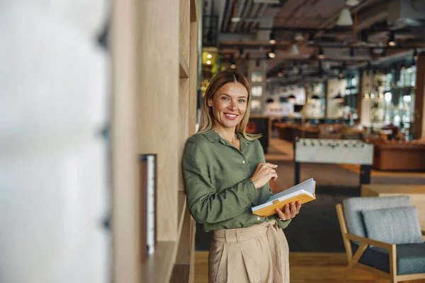 Smiling woman manager making notes in note pad while standing on cozy coworking background