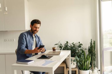 Erkek girişimci ev mutfağında dizüstü bilgisayarla çalışırken telefonu tutuyor. Mesafe çalışma kavramı