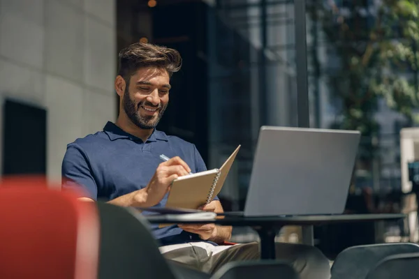 オフィスホールのラップトップで作業中にメモを作るハンサムな笑顔のビジネスマン 高品質の写真 — ストック写真