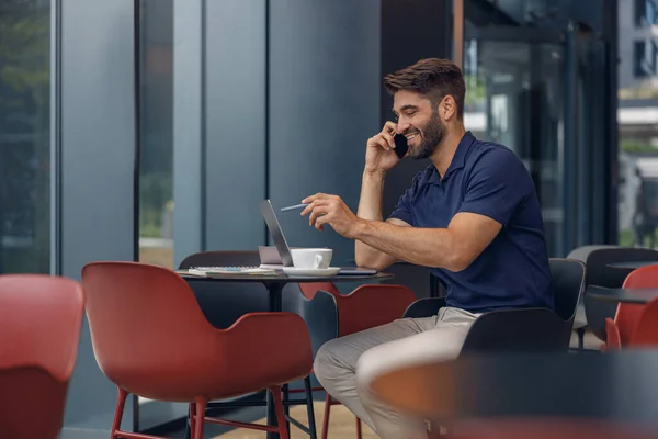 一位英俊的男性自由职业者一边在同事的笔记本电脑上工作 一边与客户通电话 — 图库照片