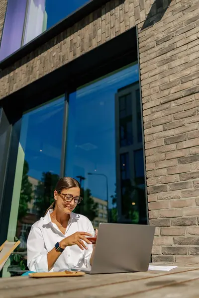 漂亮的女商人坐在咖啡厅的阳台上喝咖啡 还在笔记本电脑上工作 — 图库照片