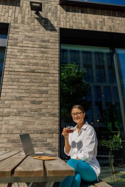 Gülümseyen bayan girişimci kahve içiyor ofis terasında dizüstü bilgisayarda çalışıyor. 