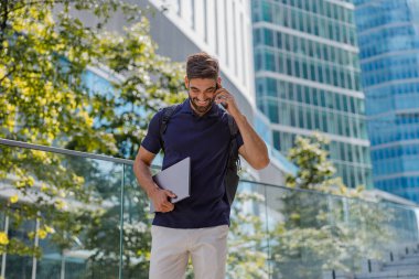 Gülümseyen iş adamı, gökdelenlerin arkasında dizüstü bilgisayarla bekleyen müşterisiyle telefonda konuşuyor.