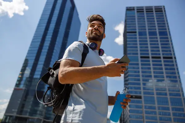 Man with phone and bottle of water standing on skyscrapers background after training and looks away