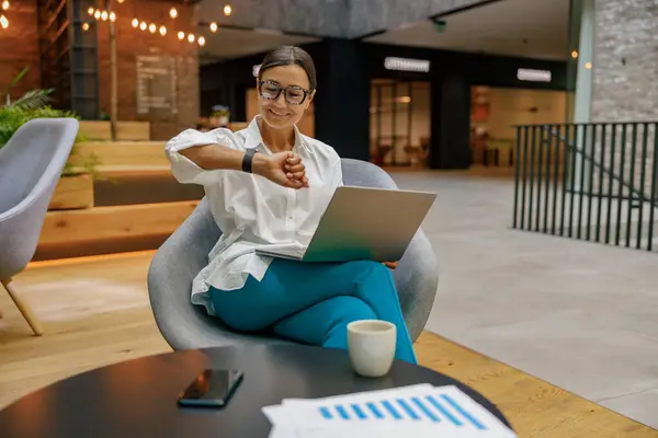 在现代办公背景下 女经理一边用手表看时间 一边用笔记本电脑 — 图库照片