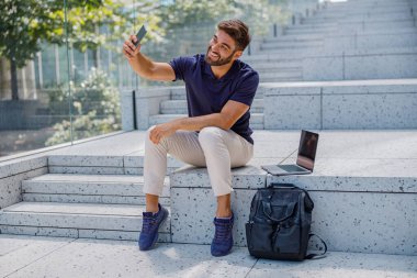 Telefonla selfie çeken yakışıklı adam modern şehir arka planında merdivenlerde oturuyor.
