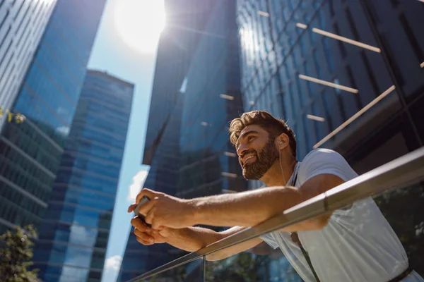 Smiling man in casual clothes standing on skyscrapers background and listen music in headphones