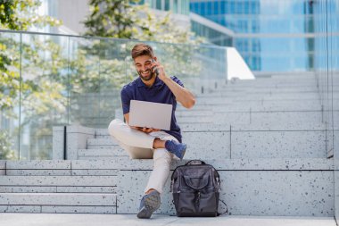 Gülümseyen işadamı müşterisiyle telefonda konuşuyor. Dizüstü bilgisayarla gökdelenlerin arkasında oturuyor.