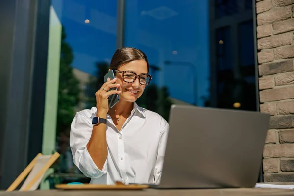 在办公室平台上的笔记本电脑上一边对着眼镜打电话一边笑着的女企业家 — 图库照片