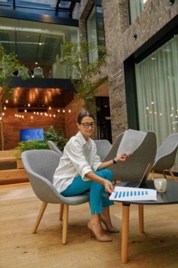 Gülümseyen stil sahibi iş kadını dizüstü bilgisayarla modern ofis arka planında oturuyor. 