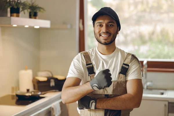 Smiling handsome handyman in uniform looking at camera and showing sign Ok standing in kitchen