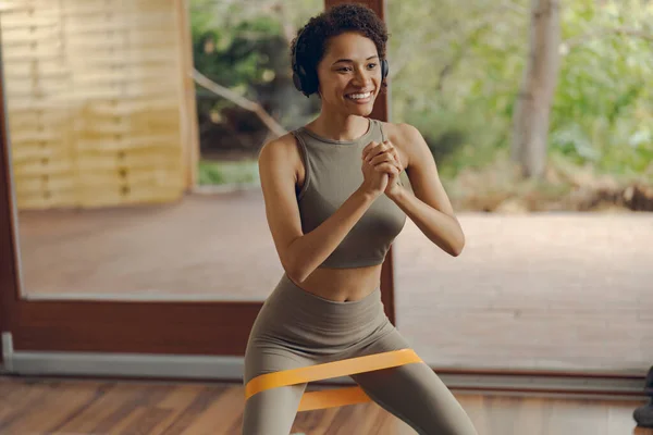 stock image Smiling woman in activewear doing squats with fitness elastic band on mat at home