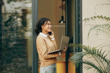 Serbest çalışan kadın, evden terasta ayakta duran dizüstü bilgisayarla telefonla konuşuyor.