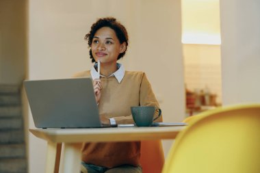 Dizüstü bilgisayarda çalışan serbest çalışan bir kadın uzaktan evde oturuyor ve başka tarafa bakıyor. Mesafe çalışma kavramı