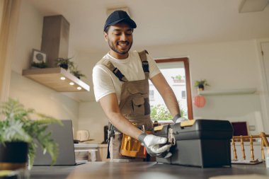 Üniformalı olumlu bir tamirci ev mutfağında duruyor ve alet çantasını açıyor.