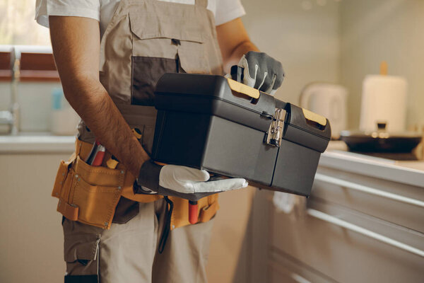 Close up of repairman in uniform standing on home kitchen and holding his tool bag