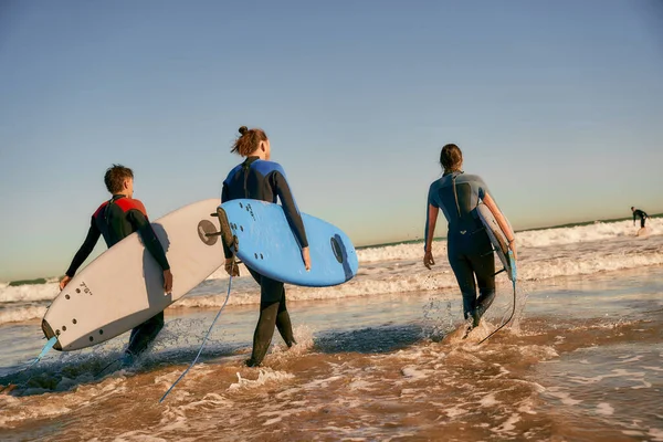 Back view of group of friends with surfboards in wetsuit entering towards ocean for surfing on waves