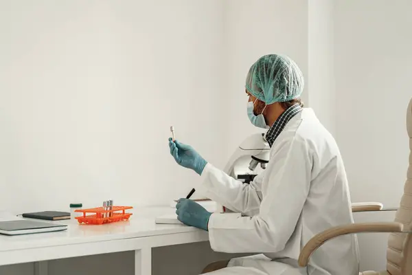 Man scientist in protective mask making notes in report sitting at laboratory with microscope