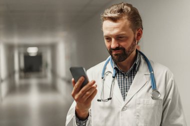 Beyaz cüppeli, steteskoplu bir erkek doktor koridor kliniğinde duruyor ve telefon kullanıyor.