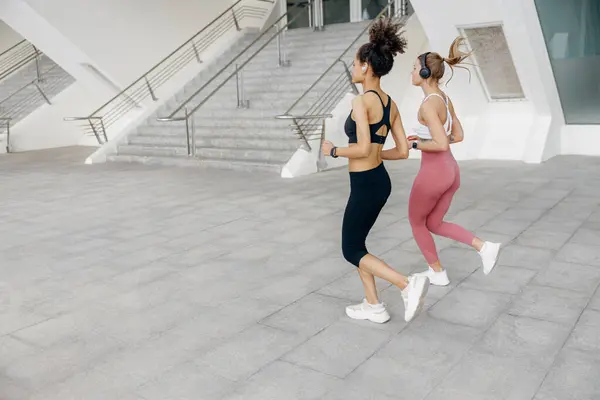 两名活跃的女运动员在现代建筑背景下沿着室外跑道并排跑 — 图库照片