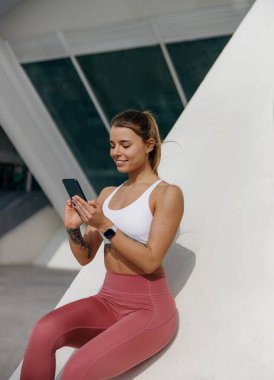 Spor kıyafetleriyle gülümseyen kadın antrenmandan sonra dışarıda oturuyor ve cep telefonu kullanıyor.