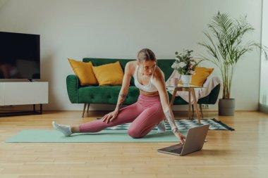 Yoga minderinde yoga yapan bir kadın rahat bir iç dizaynla dizüstü bilgisayarın önünde. 