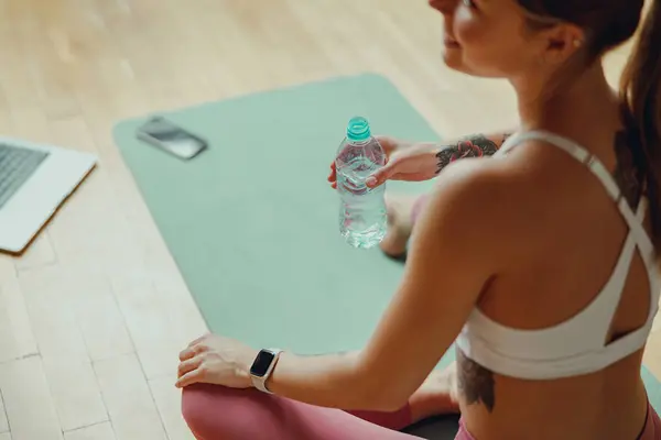 一个穿着运动服的女人正在悠闲地坐在瑜伽垫上 一只手拿着一瓶水 — 图库照片