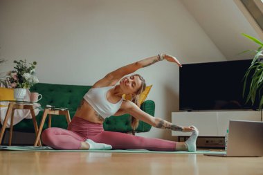 Bir kadın, oturma odasındaki ahşap döşemeli yoga minderinin üzerinde bacaklarını rahatça açıyor.