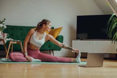 Bir kadın ahşap zeminde yoga minderinde oturuyor ve bacaklarını esnetiyor. Yüksek kalite fotoğraf