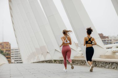 Modern binaların arka planında açık hava pistinde yan yana koşan iki aktif bayan sporcu.