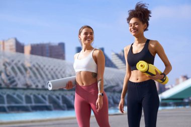 İki kadın yan yana duruyorlar, gülümsüyorlar ve yoga minderleri tutuyorlar.